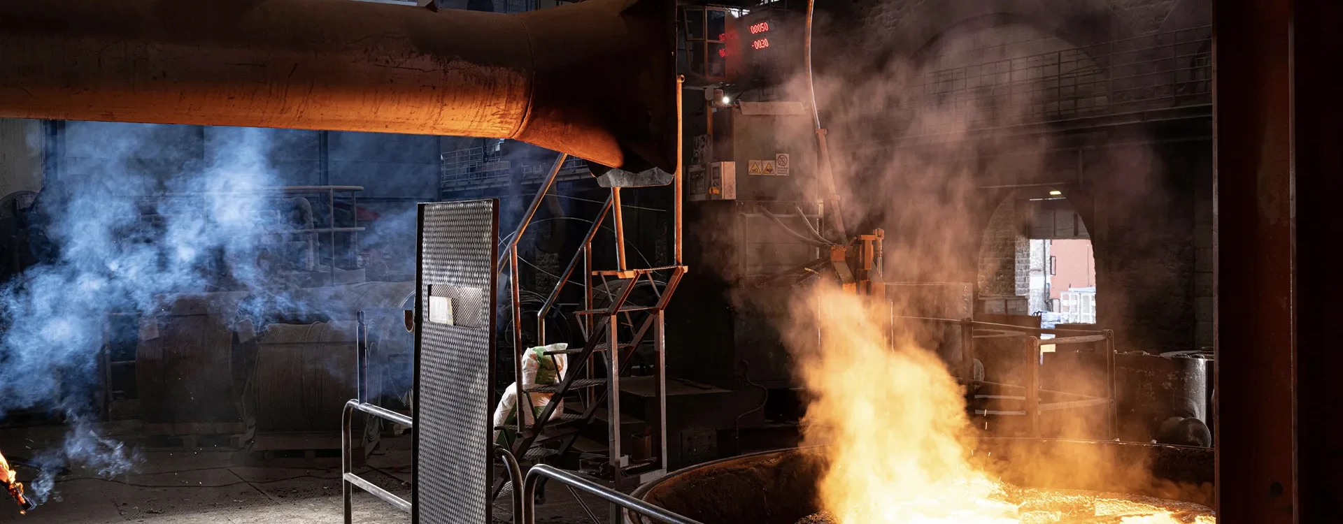 Interno di una fonderia con fiamme e denso fumo che si sprigionano da un forno fusorio, mentre un operaio in tuta protettiva supervisiona la lavorazione sullo sfondo.