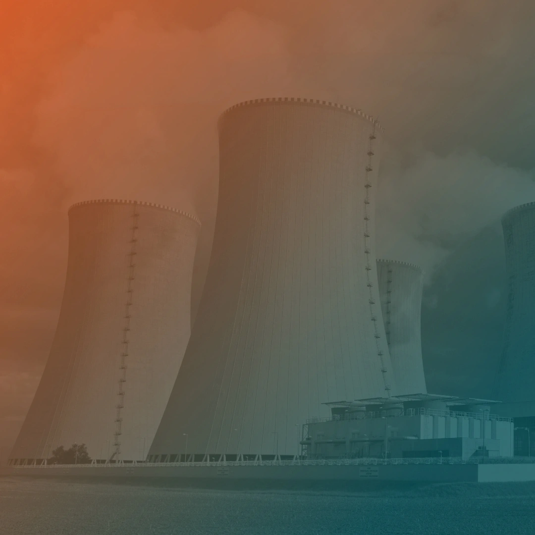 Quattro torri di raffreddamento di una centrale nucleare, con vapore che fuoriesce dalla sommità, su sfondo di cielo nuvoloso. In primo piano un campo arato e un edificio industriale con strutture blu.